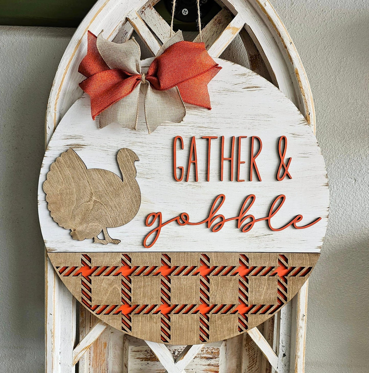 A round decorative piece with a turkey design and the text 'GATHER & GOBBLE' on a wooden surface, adorned with a bow and autumn-colored ribbons.