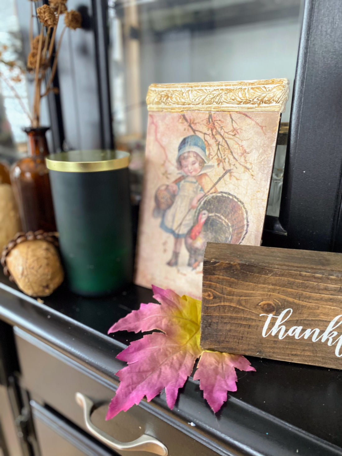 Cottage vignette with green pillar candle, vintage Thanksgiving art, wooden thankful sign, and autumn leaves.