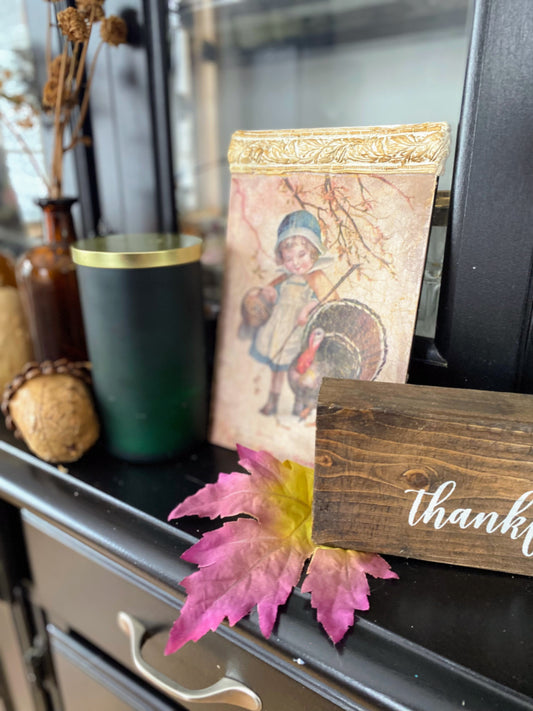 Cottage vignette with green pillar candle, vintage Thanksgiving art, wooden thankful sign, and autumn leaves.